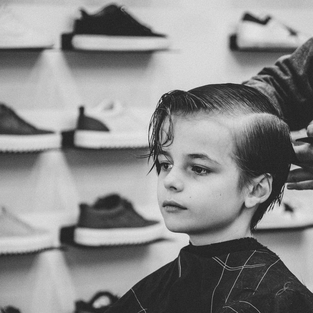 Crno-bijela fotografija: dječak u barberskoj stolici dok mu barber češlja mokru kosu