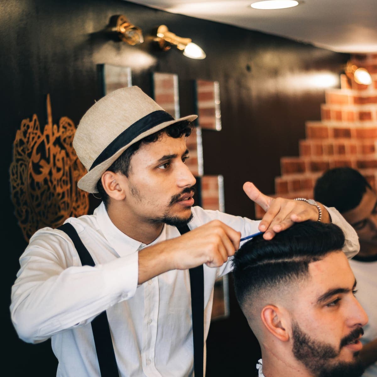 Barber češljem oblikuje svježe ošišan fade s razdjeljkom, gost s bradom sjedi u stolici