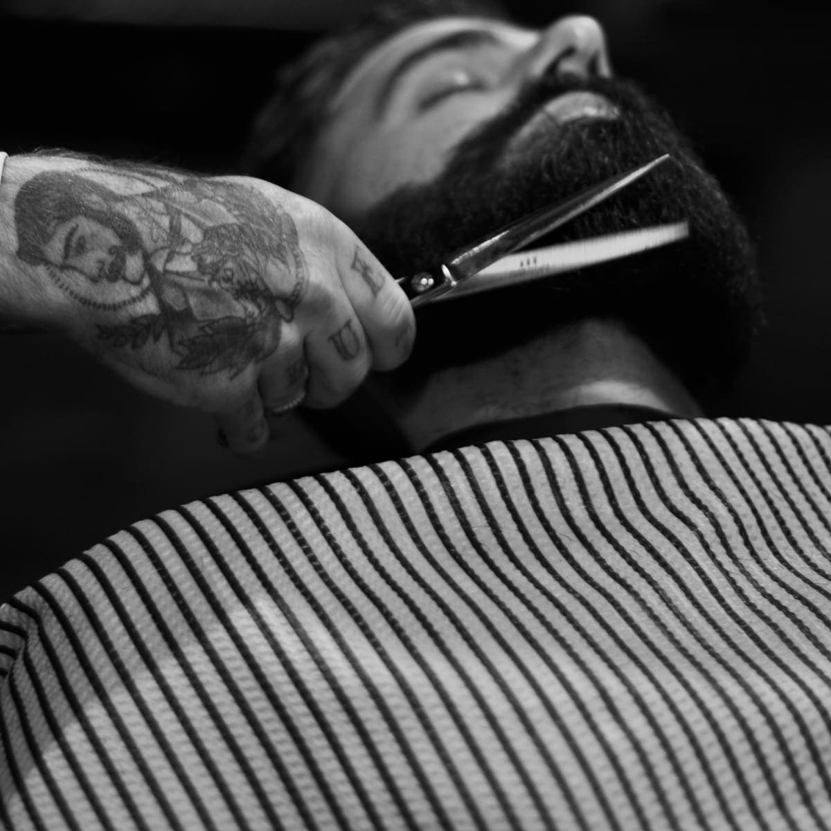 Crno-bijela fotografija: barber škarama skraćuje bradu gostu zabačene glave
