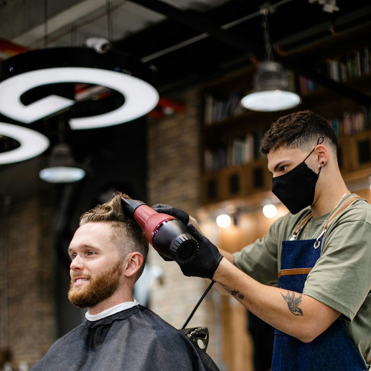 Barber fenom oblikuje kosu nasmijanom gostu s bradom, u salonu s ciglenim zidom